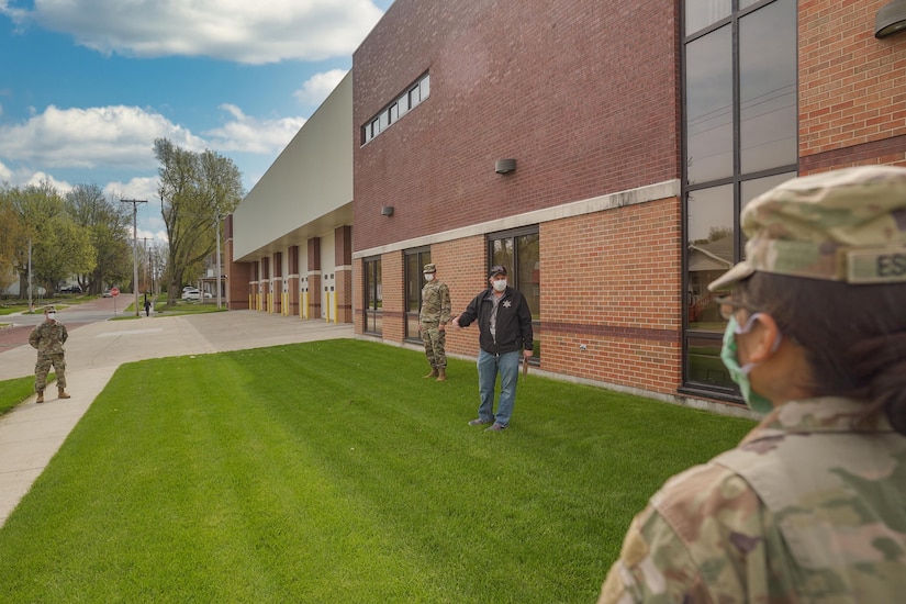 Civilians and military personnel wearing face masks stand on the lawn and sidewalk in front of a municipal building.