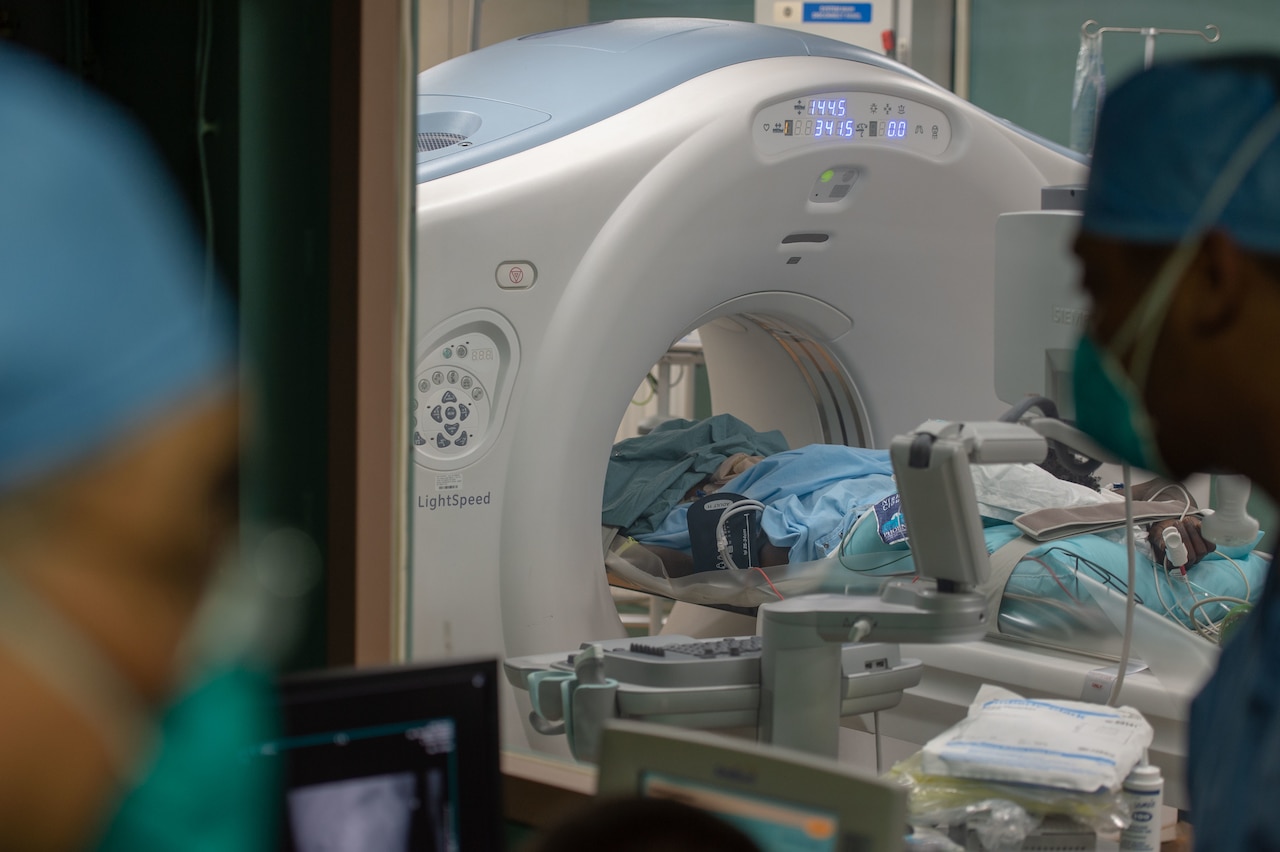 A patient undergoes a CT scan as two sailors wearing masks monitor the test.