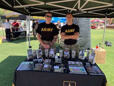 White female in black and yellow army shirt and green camouflage pants stands with a white male in black and yellow Army t-shirt in green camouflage pants behind a table of print brochures under a tent.