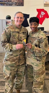 white female in green camouflage uniform and black female wearing camouflage uniform hold two sides of a business card that depicts Rosie the Riveter.