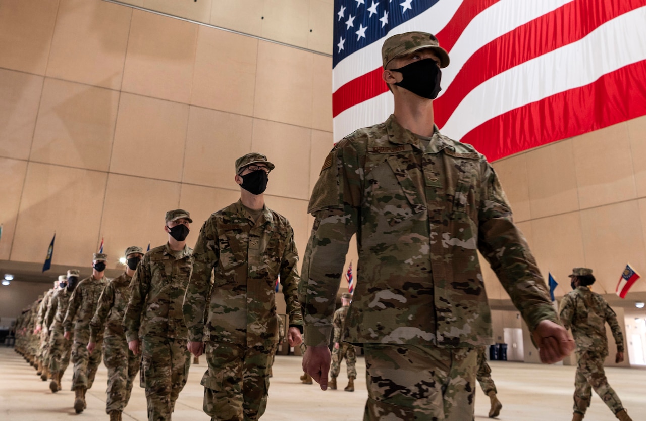 Multiple service members wearing black face masks march in a line. An American flag hangs above them.