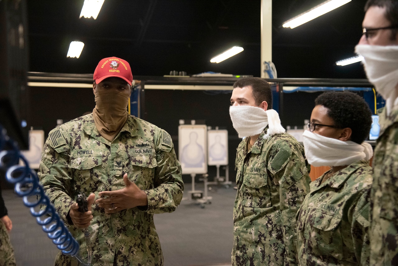 A man in military uniform and wearing a red hat talks with young military members who are standing near him. The man holds a pistol that is attached to an air hose. Everybody wears a face mask.