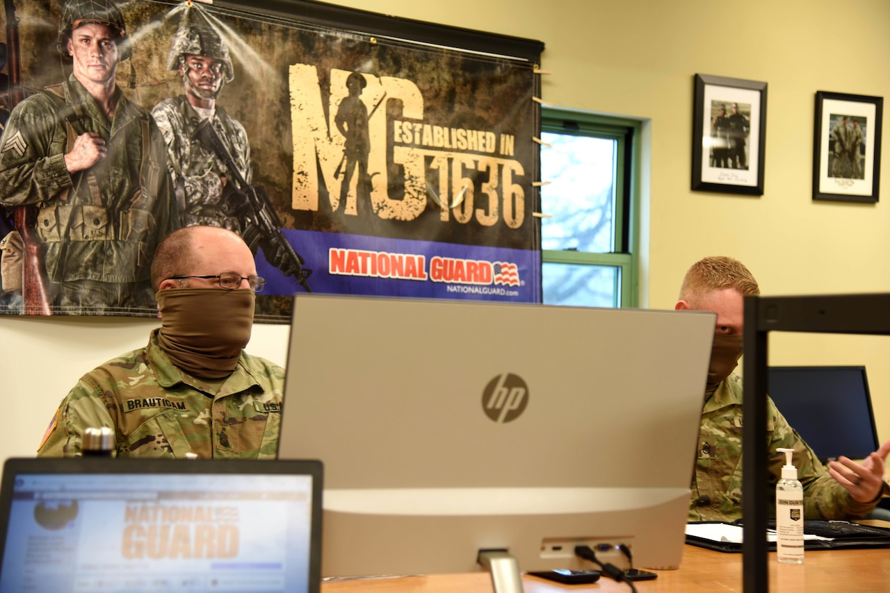 Two military members sit behind a computer in an office setting. Behind them is a banner advertising the National Guard.