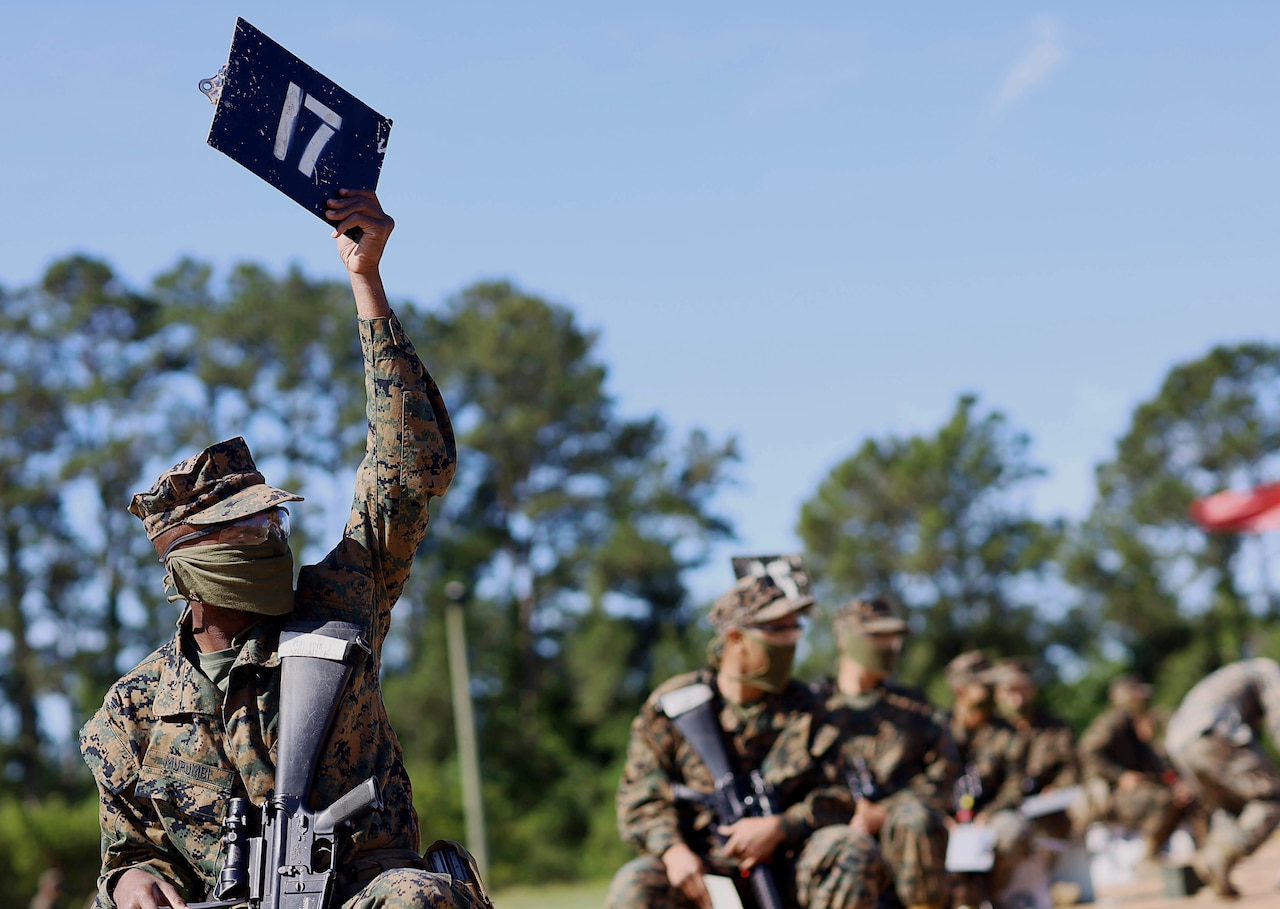 Multiple service members in camouflage uniforms carry rifles. One holds a sign with the number 17 on it.