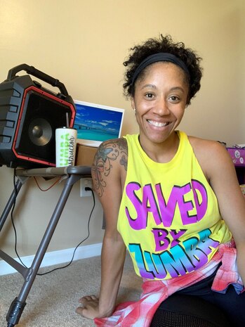 Photo shows a woman sitting in front of her tablet and music speaker smiling for the camera with the words, "Saved by Zumba" on her workout shirt.