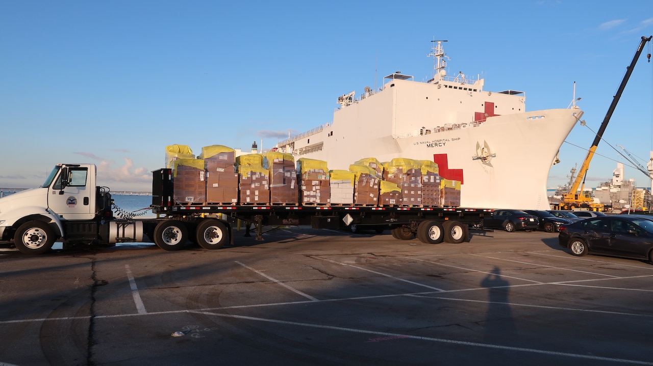 A flatbed truck loaded with pallets of supplies sits on the dock near a white hospital ship.