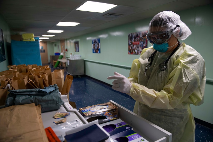 A woman puts on personal protective equipment near a table that holds dozens of paper bags.