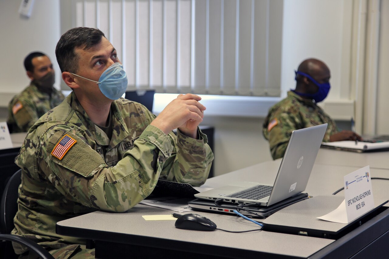 A soldier wearing a face mask sits at a laptop computer.