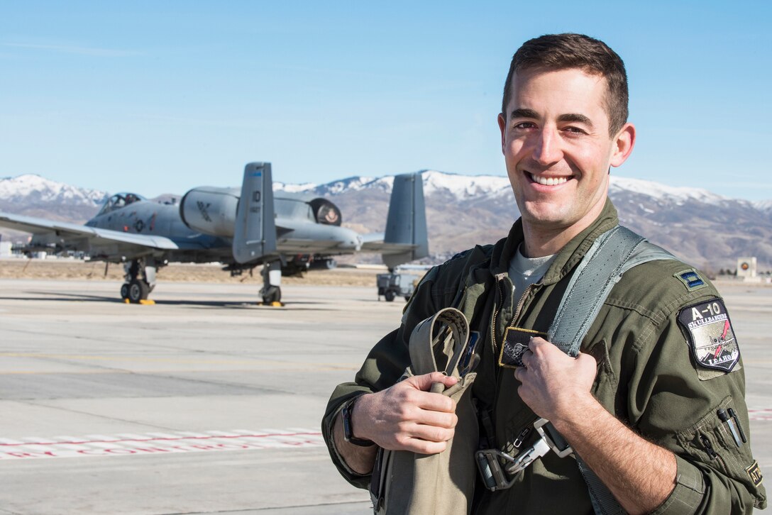 An A-10 pilot wearing a flight suit poses for a photo with an A-10 aircraft on the tarmac behind him and mountains in the background.