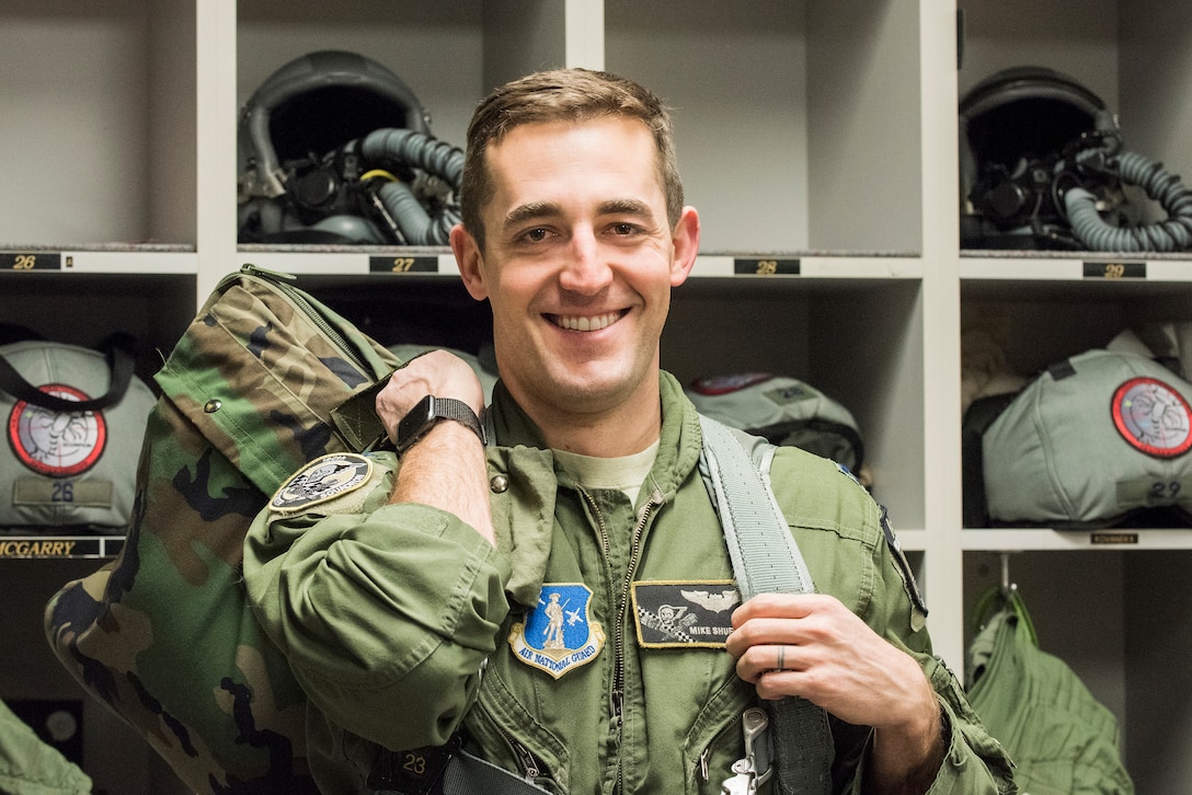 A pilot wearing a flight suit and harness, carrying a bag slung over his shoulder, poses for the camera. A wall of storage cubes with equipment is in the background.