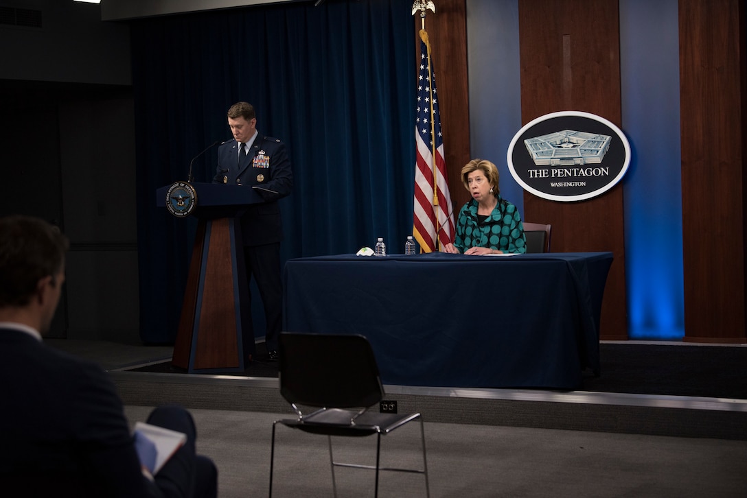 A woman sits at a desk as a man stands beside her at a podium in front of an audience.