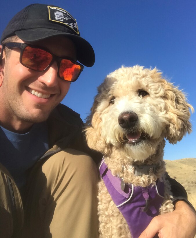 A man in sunglasses and ball cap kneels to take a selfie with his fluffy golden-colored dog.