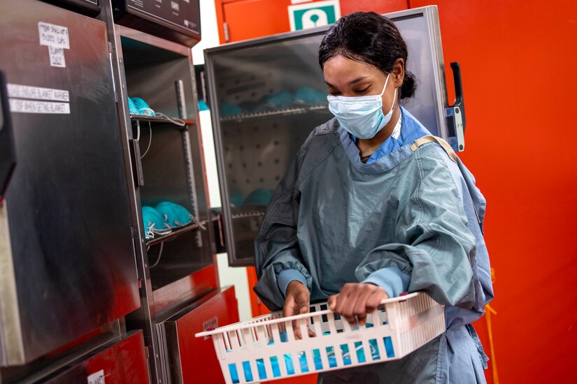 A woman in hospital scrubs holds a basket full of paper masks that she is placing in a large metal cabinet.