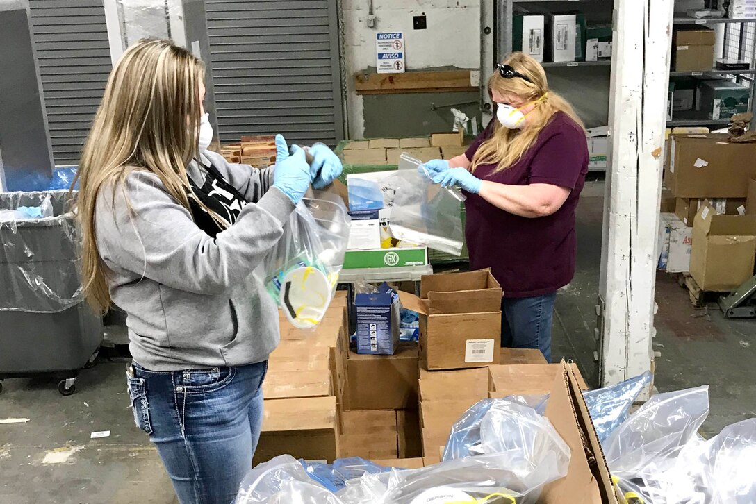 Two women put masks into plastic bags. In the foreground are cardboard boxes filled with individually wrapped masks.