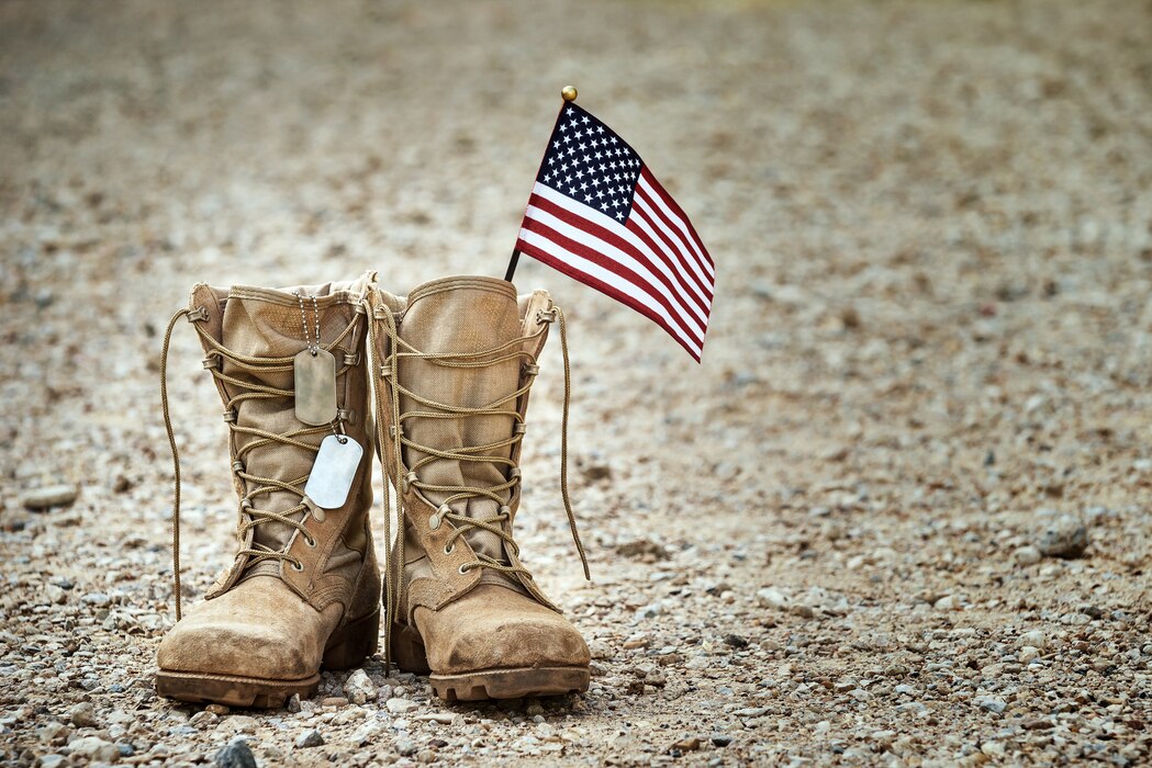 photo shows a pair of combat boots with dog tags hanging off them and a flag sticking up out of the boot.