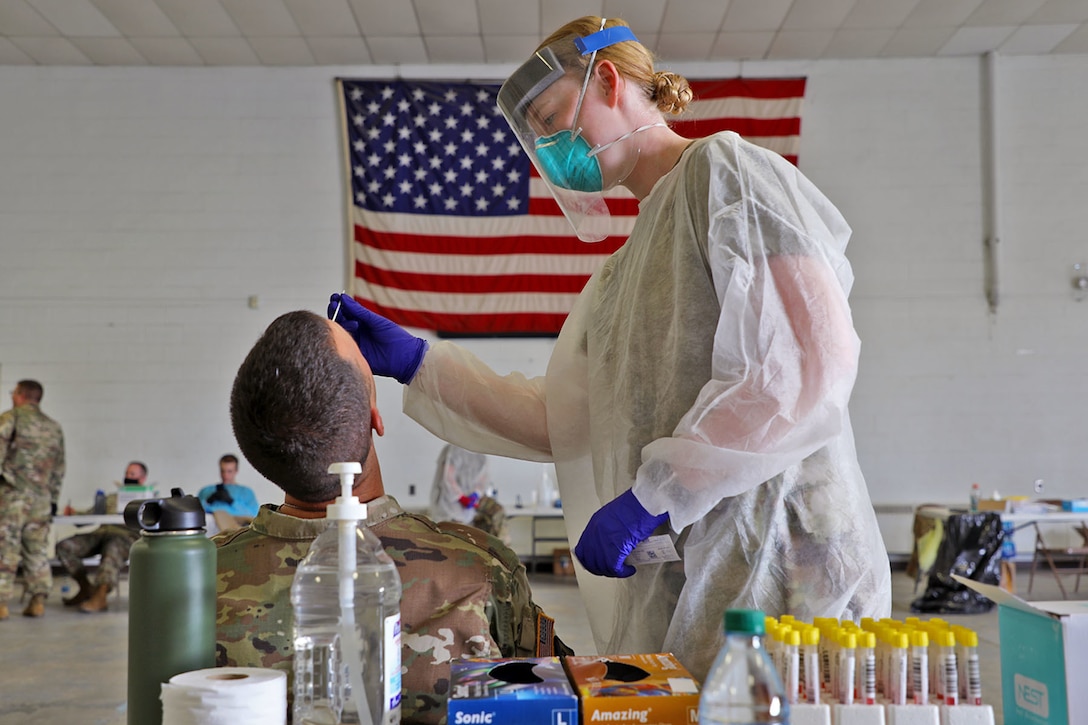 Female in white gown and blue gloves, clear face shield inserts a tester into a male soldier's nose to test for the coronavirus.