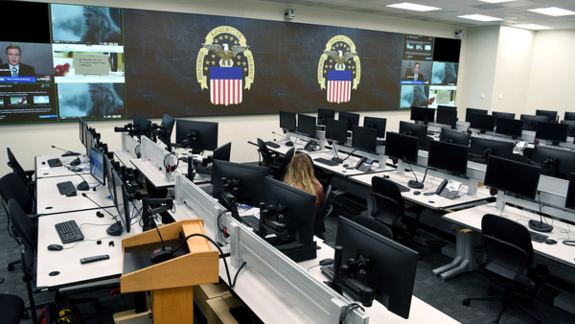 Room with computers on white desks for about 20 people. Large TV screens in the front of the room.