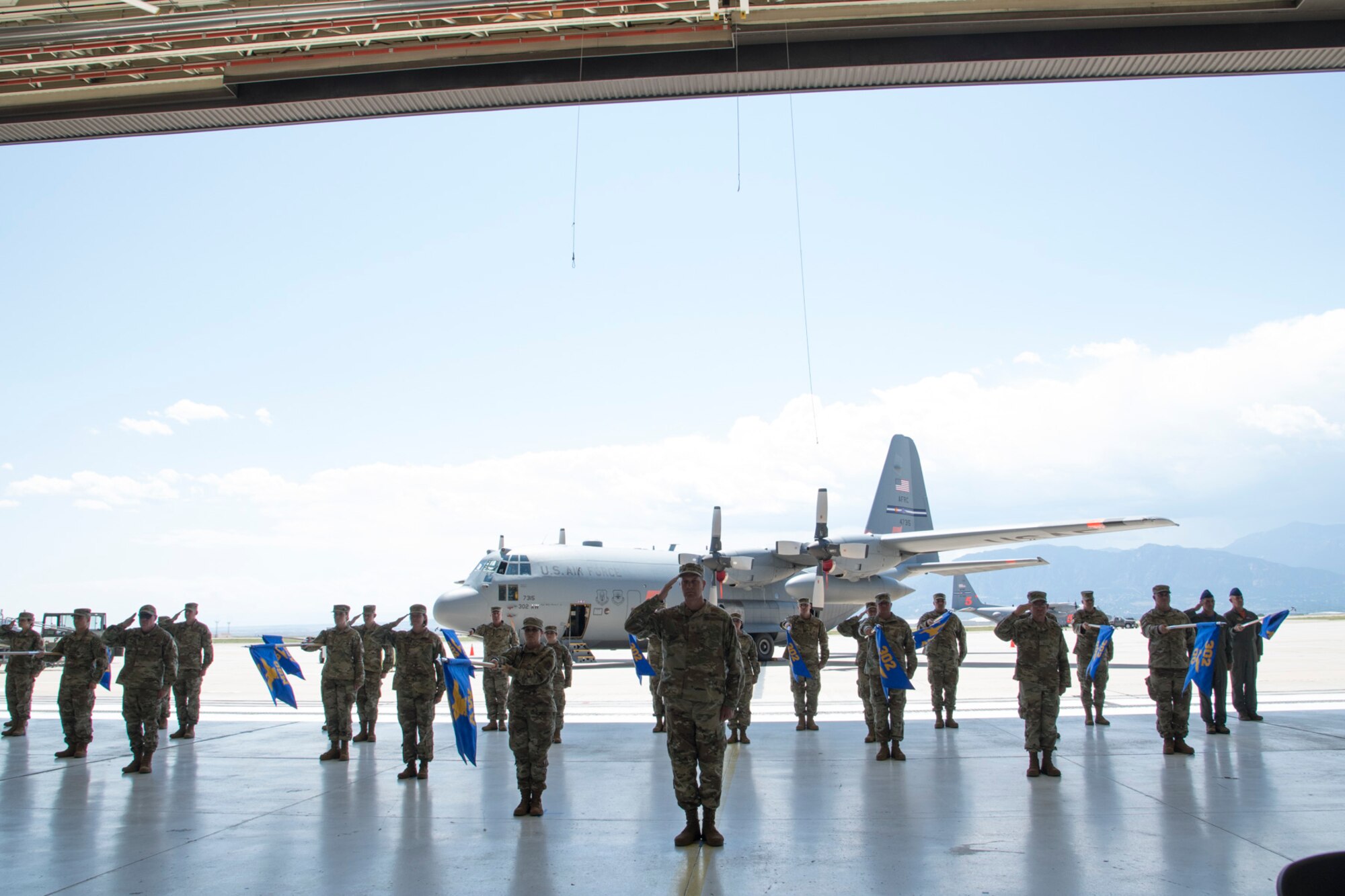 Members of the 302nd Airlift Wing salute as a formation in a aircraft hangar with a C-130 Hercules behind them.