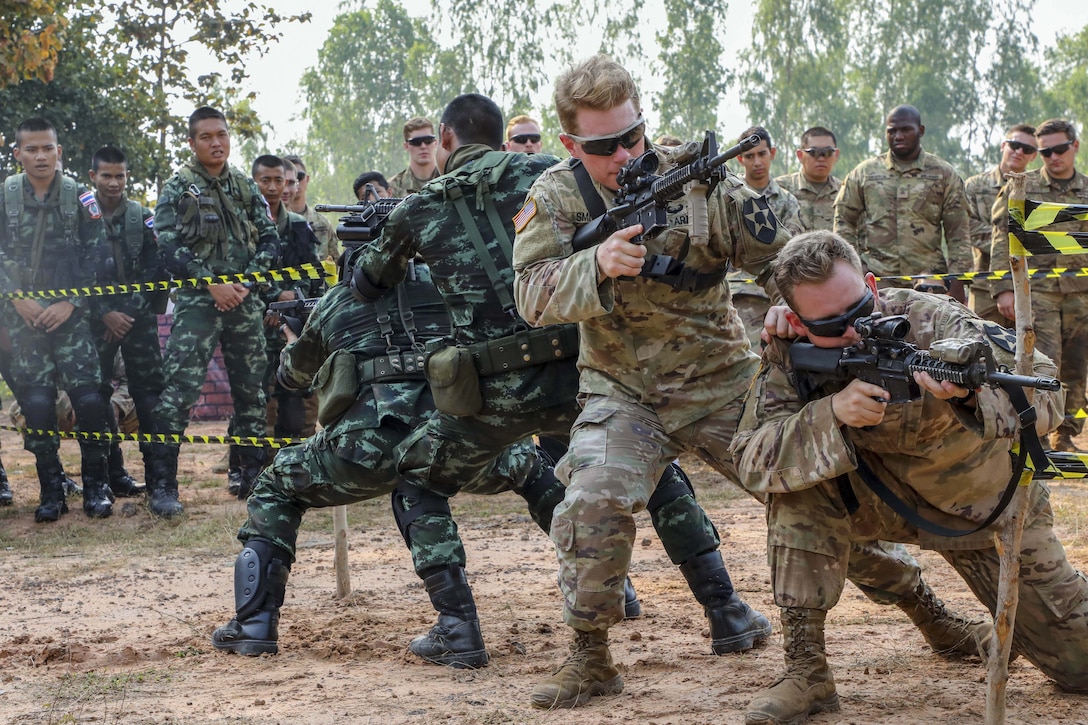 Two U.S. soldiers and two Thai soldiers crouch and aim rifles in opposite directions as other soldiers watch during a training event.