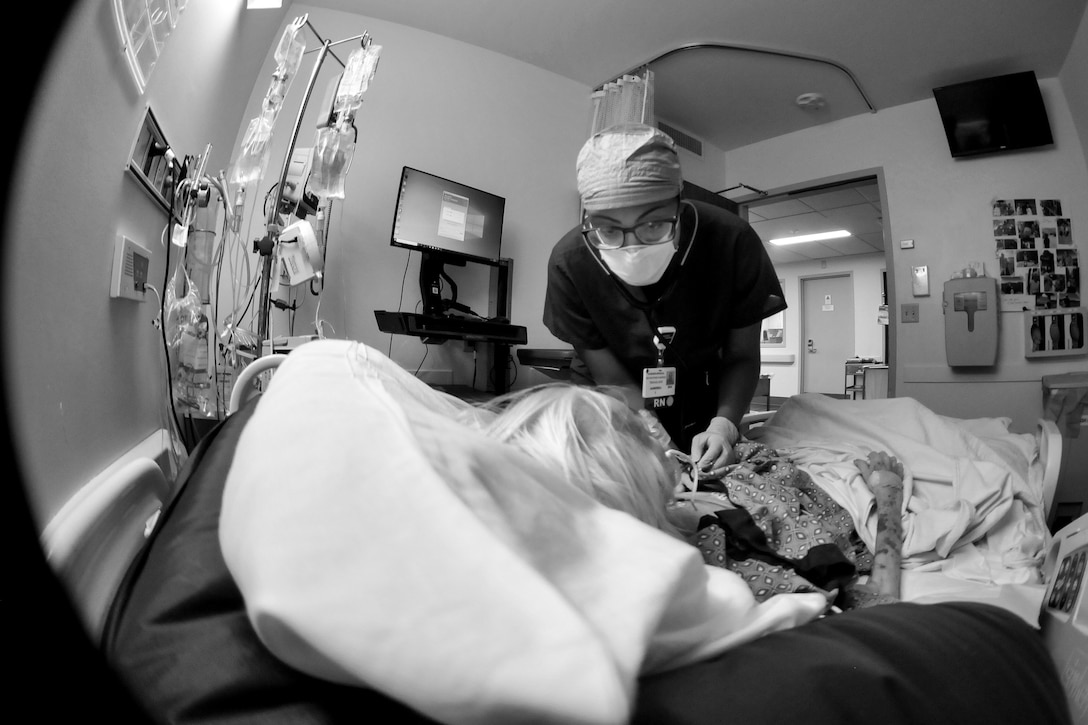 An airman wearing a face mask leans over a patient in a hospital bed.