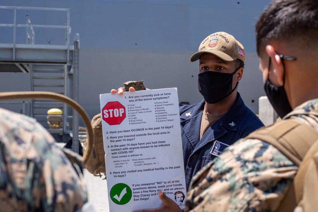 A sailor wearing a face mask holds a sign describing the symptoms of COVID-19.