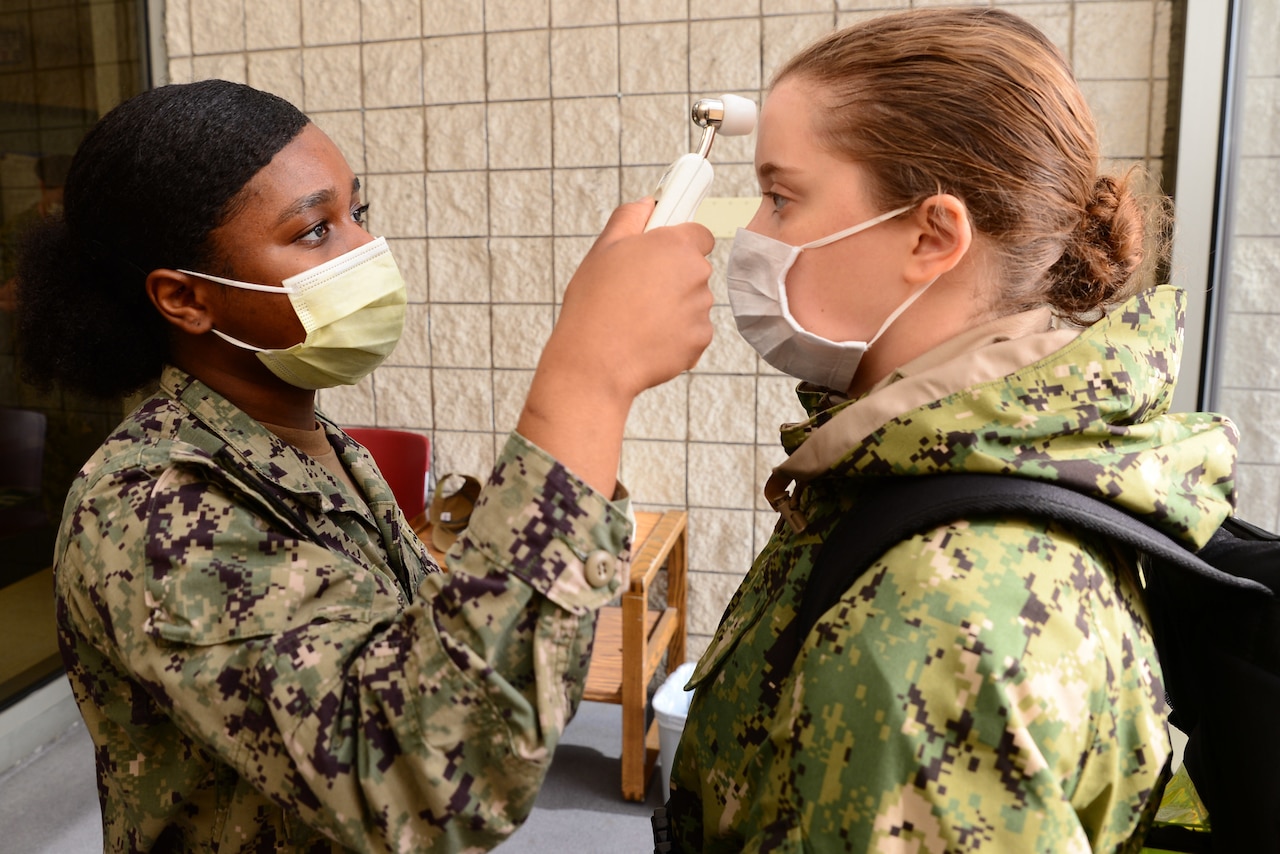 A woman dressed in a camouflage uniform and wearing a face mask puts a thermometer near the forehead of another woman wearing a face mask and fatigues.