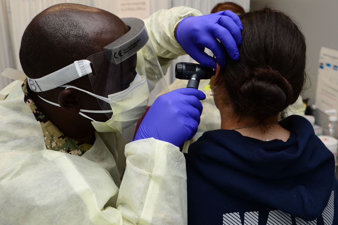 A man wearing personal protective equipment uses a medical instrument to look into the ear of a woman dressed in a camouflage uniform.