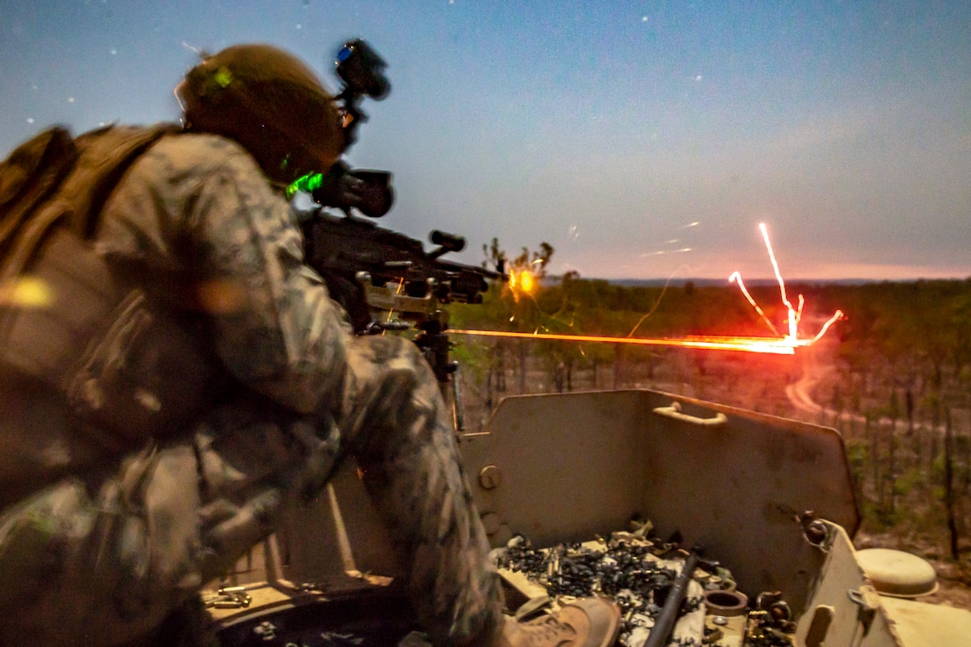 A line of red light shoots into a field as a Marine fires a weapon in low-light conditions.