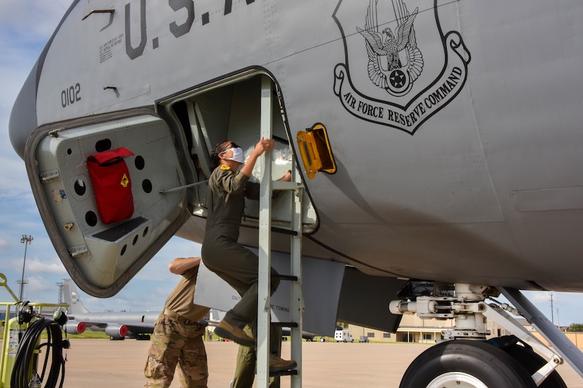 A pilot wearing sunglasses and a face mask climbs a ladder that leads into the cockpit of a tanker aircraft.