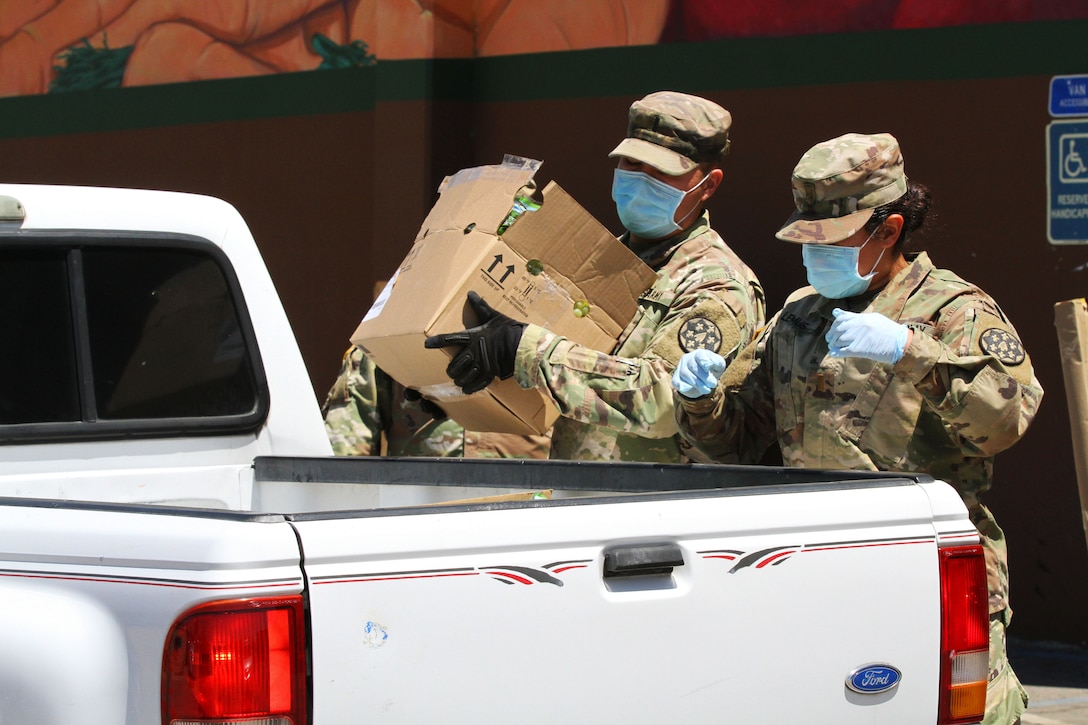 Two soldiers wearing face masks and gloves load boxes of food into the back of a vehicle.