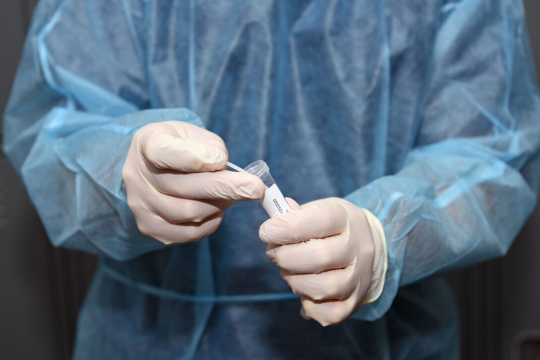 A soldier wearing personal protective equipment holds a nasopharyngeal swab and test tube.