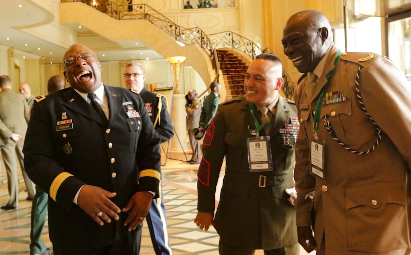 Three men in military uniforms laugh together in a large foyer.