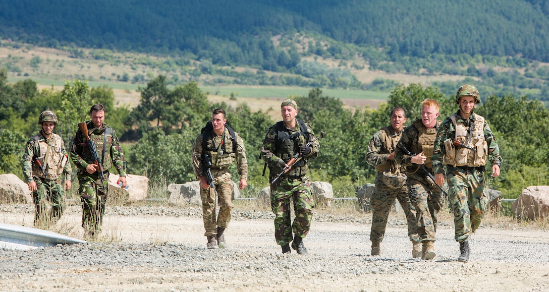 Seven uniformed personnel, many with rifles, walk together on a gravel road.
