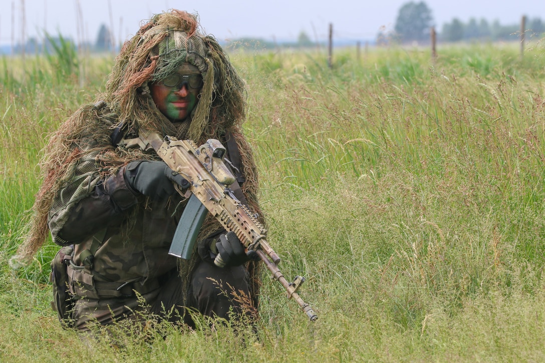 A service member kneels in a field. He holds a rifle, and wears a Guillie suit.