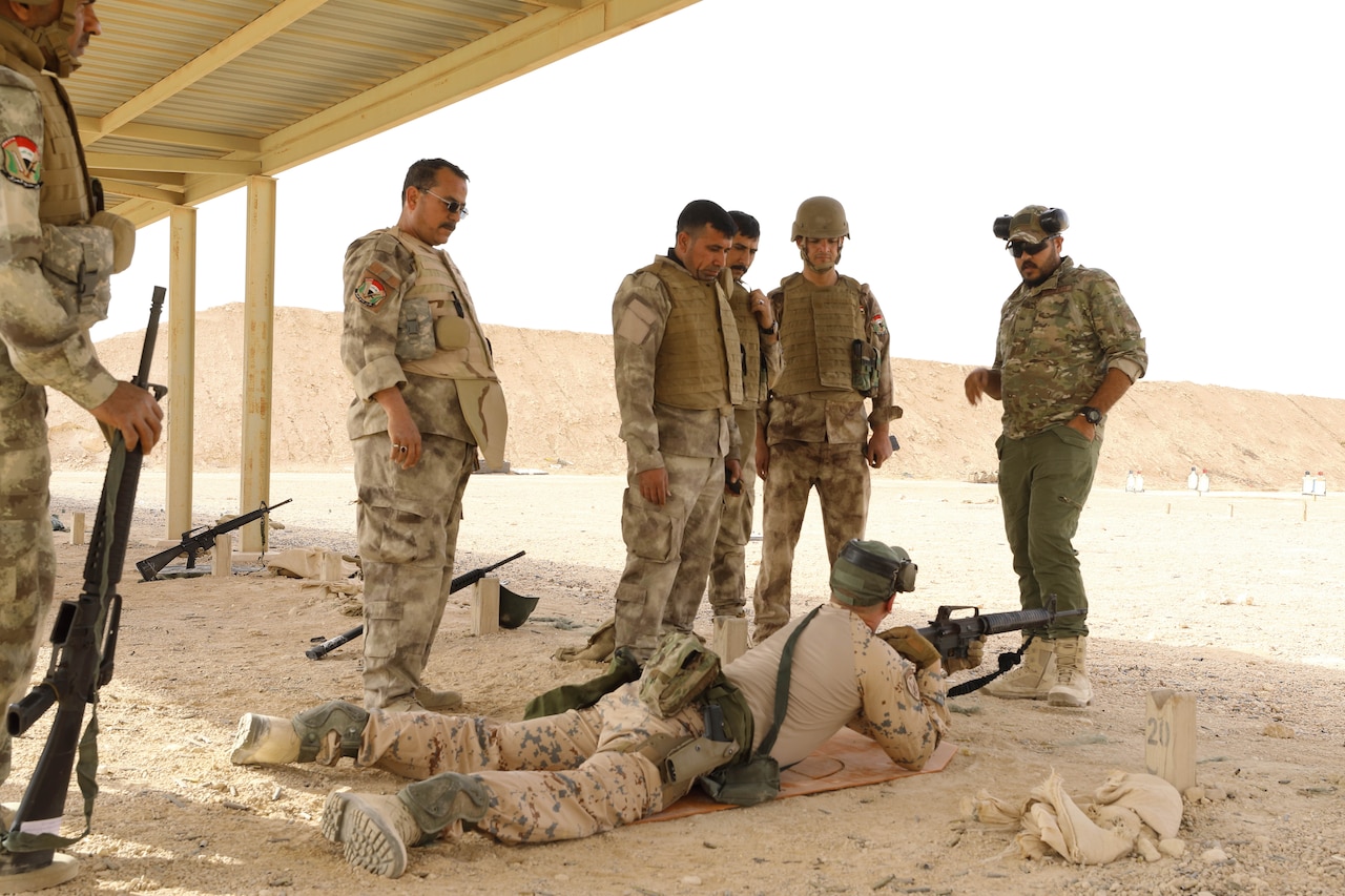 A service member lies on the ground with a rifle. Other service members are standing around him.