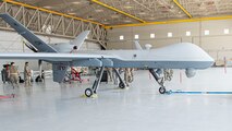 MQ-9 Reaper sits in hangar with 6 maintenance members preparing for routine maintenance.