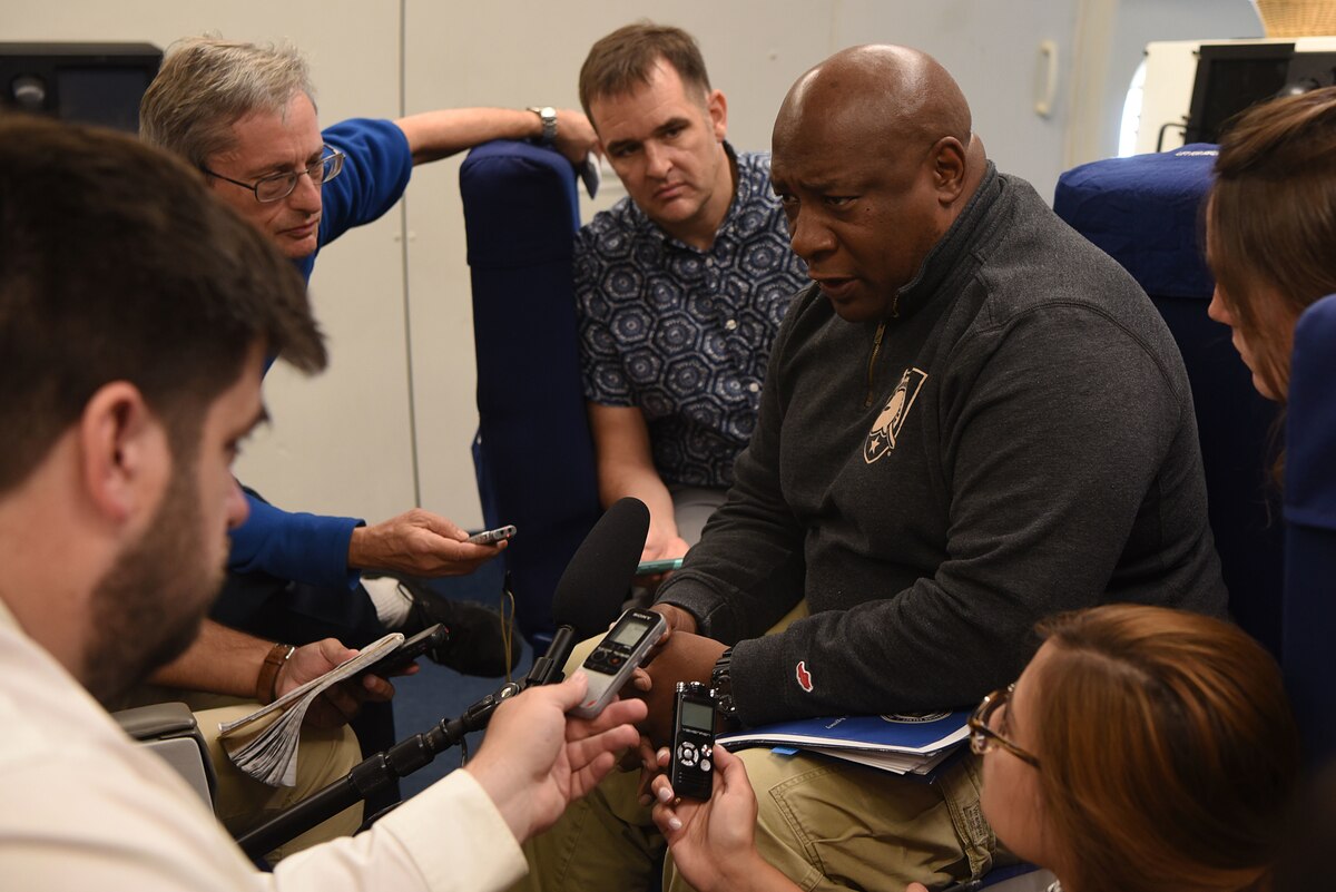 Man sits among five reporters with microphones and notepads.