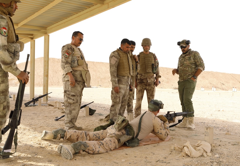 A service member lies on the ground with a rifle. Other service members are standing around him.
