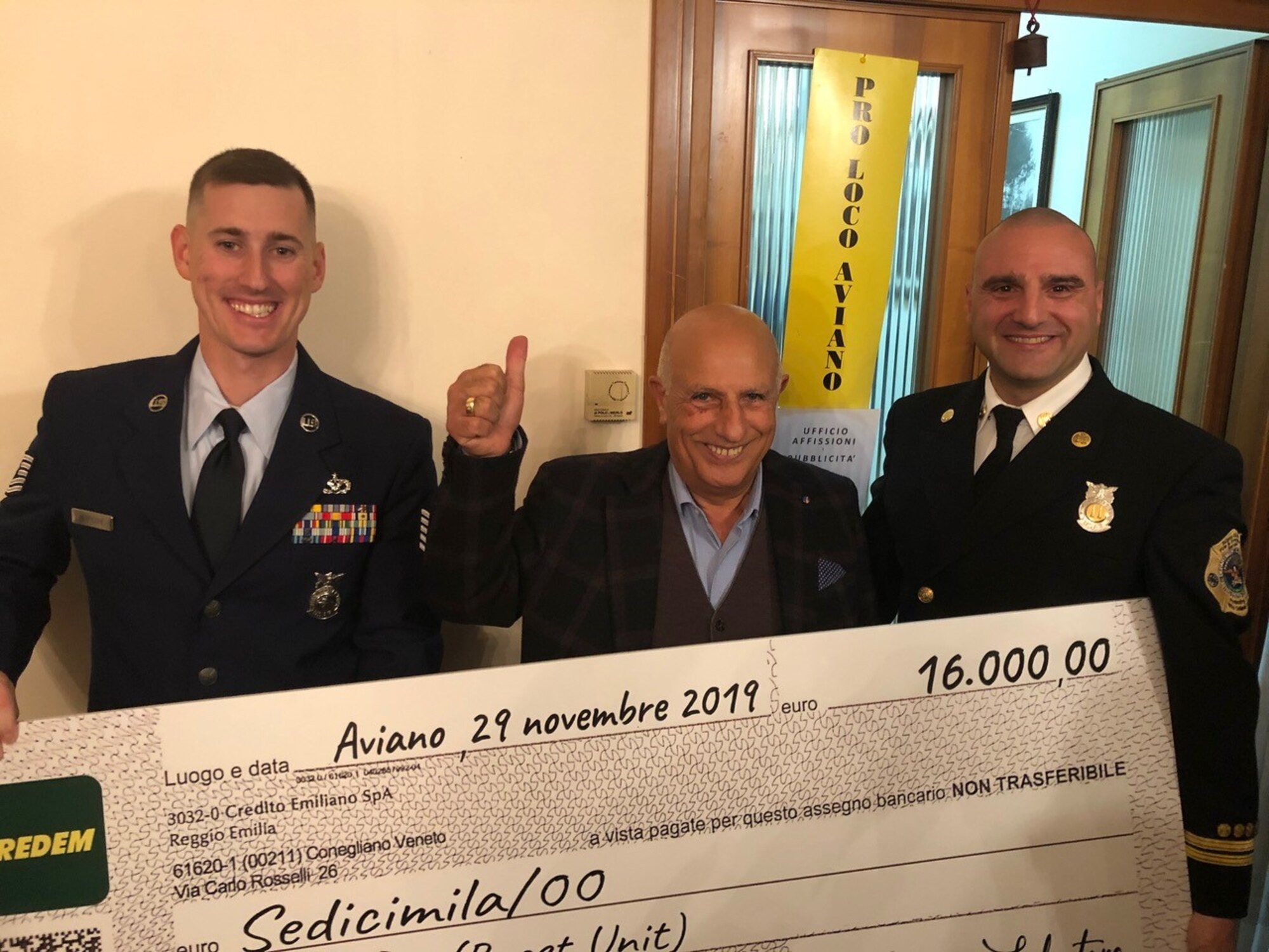 Tech. Sgt. Robert Perricone, 31st Civil Engineer Squadron assistant chief of health and safety (left), Salvatore Brunno, Volunteer Association of Aviano president (middle), and Fabrizio La Marca, 31st Civil Engineer Squadron firefighter (right), hold a check while posing for a photo in Aviano, Italy, Nov. 29, 2019. The Aviano Firefighter Association sold custom shirts and hoodies to raise money for breast cancer awareness. (Courtesy photo)