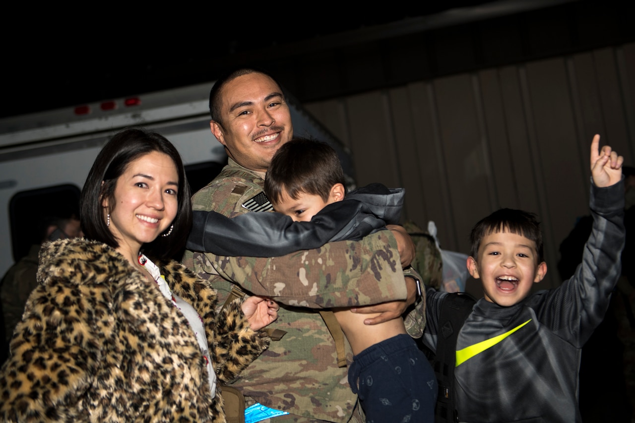 A man in a military uniform holds a small boy. Near them are another small boy and an adult woman.