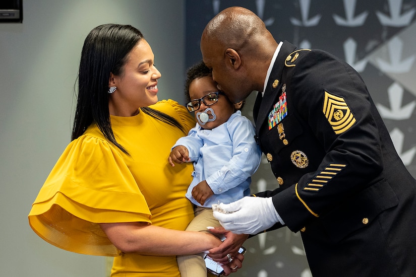 A woman holds a baby.  A man in military uniform kisses the child on the head.