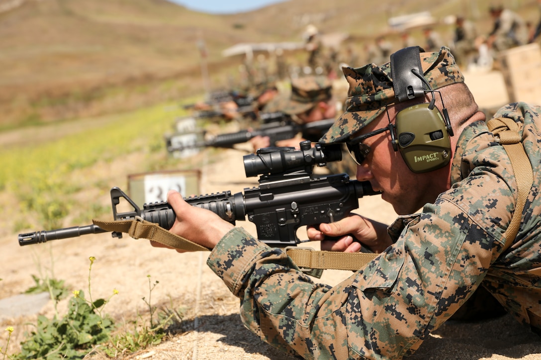 A row of service members lie on the ground and fire their rifles.