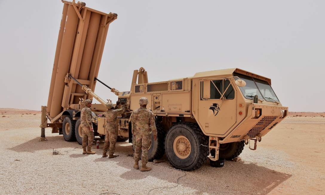 Three service members work on a large military vehicle in the desert.