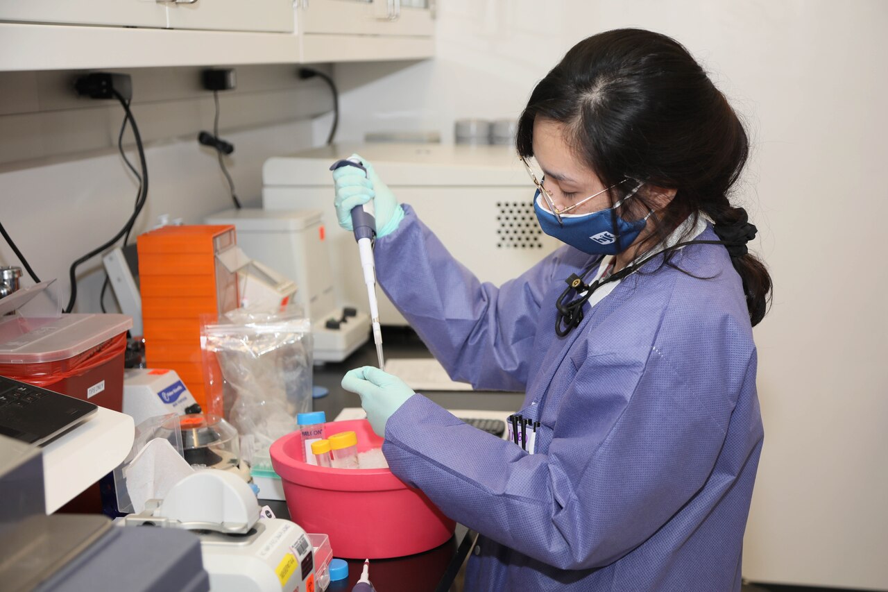 A researcher in a mask and gloves uses hand-held lab instruments at a counter.