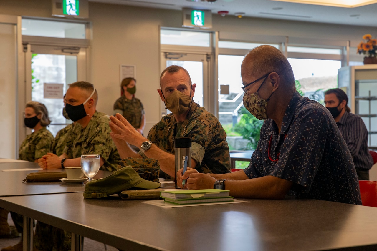 Service members and civilians talk while seated at a conference table. All are wearing face masks.