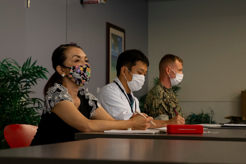 A civilian woman, a civilian man and a Marine, all wearing face masks, sit at a conference table during a meeting.