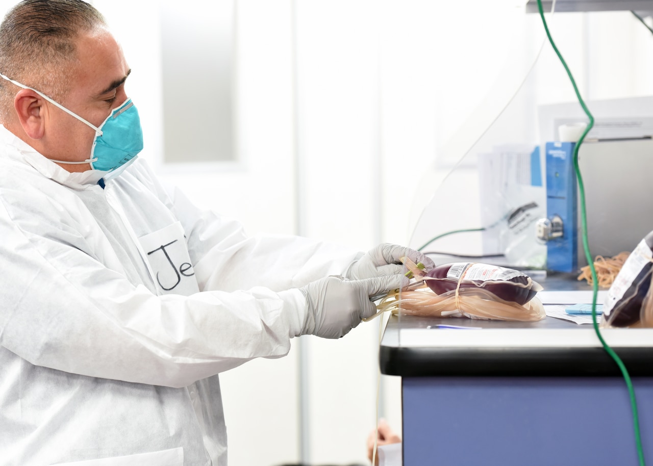 A man wearing a face mask, gloves and a white smock handles bags of donated blood.