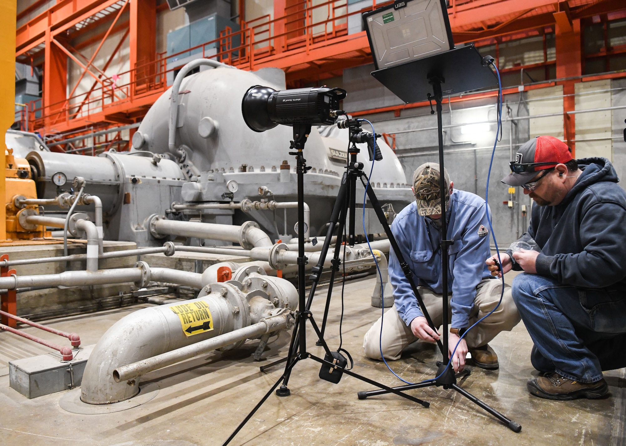 On March 4, Arnold Engineering Development Complex Condition-Based Maintenance craftsmen Nathan Dotson, right, and Landon Culver place vibration reduction pads under the feet of a tripod holding a Motion Amplification Camera, while setting up the camera to help identify the source of excessive vibrations in machinery in the C Plant of the Engine Test Facility at Arnold Air Force Base, Tenn. The vibration reduction pads help stabilize the camera to minimize the influence of vibrations through physical contact on the video being captured. (U.S. Air Force photo by Jill Pickett)