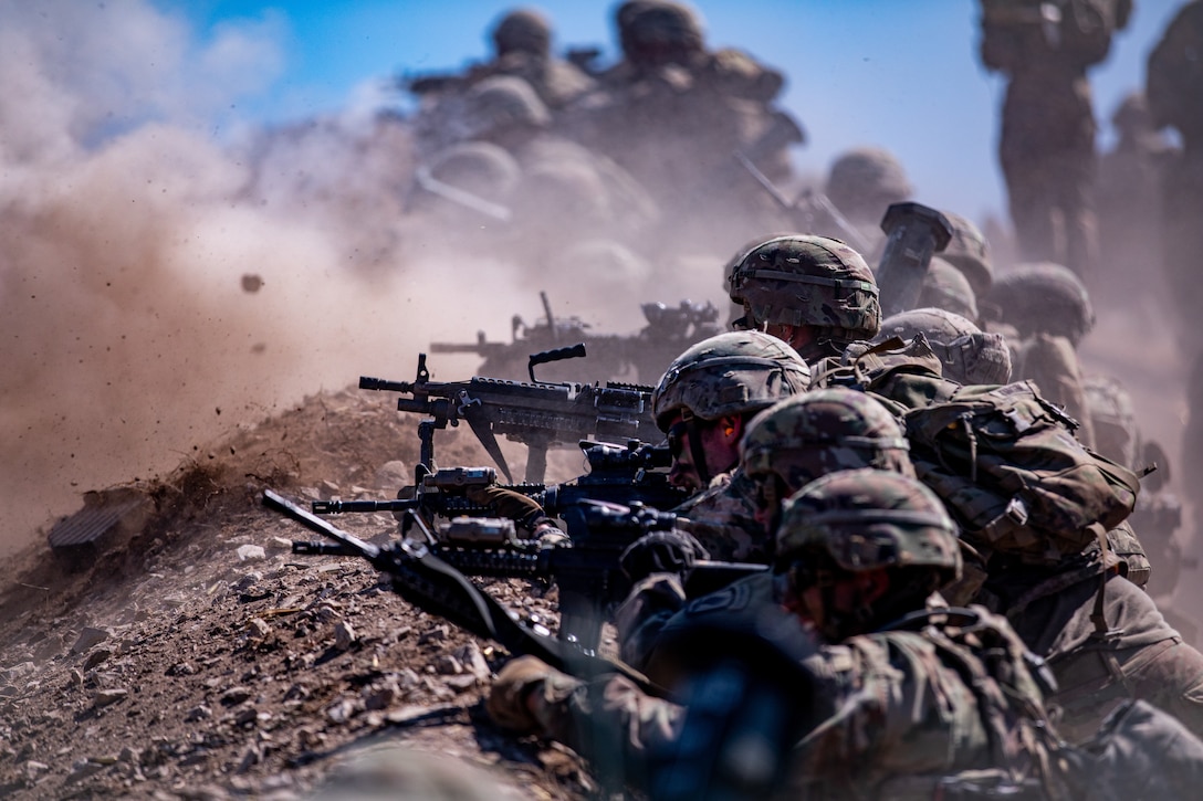 Soldiers lay in a line and aim weapons as dirt clouds rise in the background.