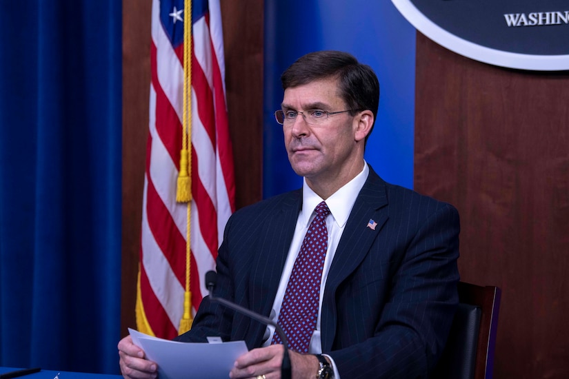 A man holding index cards sits at a table in front of a large television. Behind him are an American flag and a blue Pentagon seal.